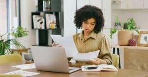 Shot of a young woman going through paperwork while working on a laptop at home